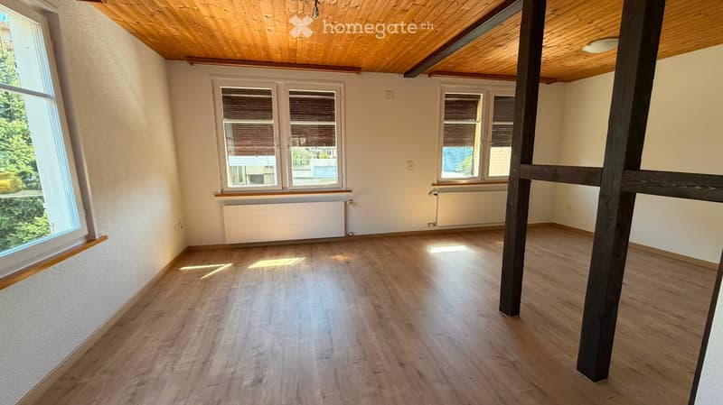 Empty room with wooden flooring, white walls, and large windows with blinds. The room features a slanted wooden ceiling and a visible support beam.