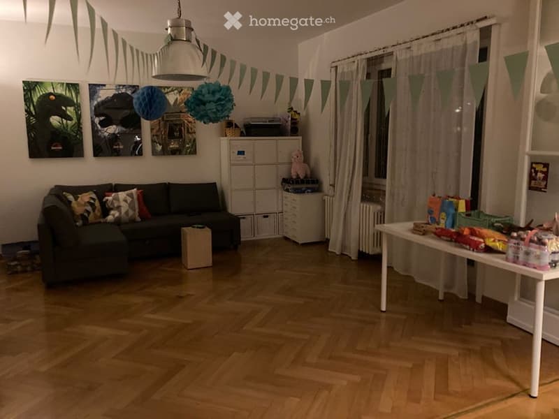 A cozy living room with a dark green sectional sofa, colorful throw pillows, a white coffee table, a storage unit with a pink stuffed animal, and a table filled with children's toys and decorations.