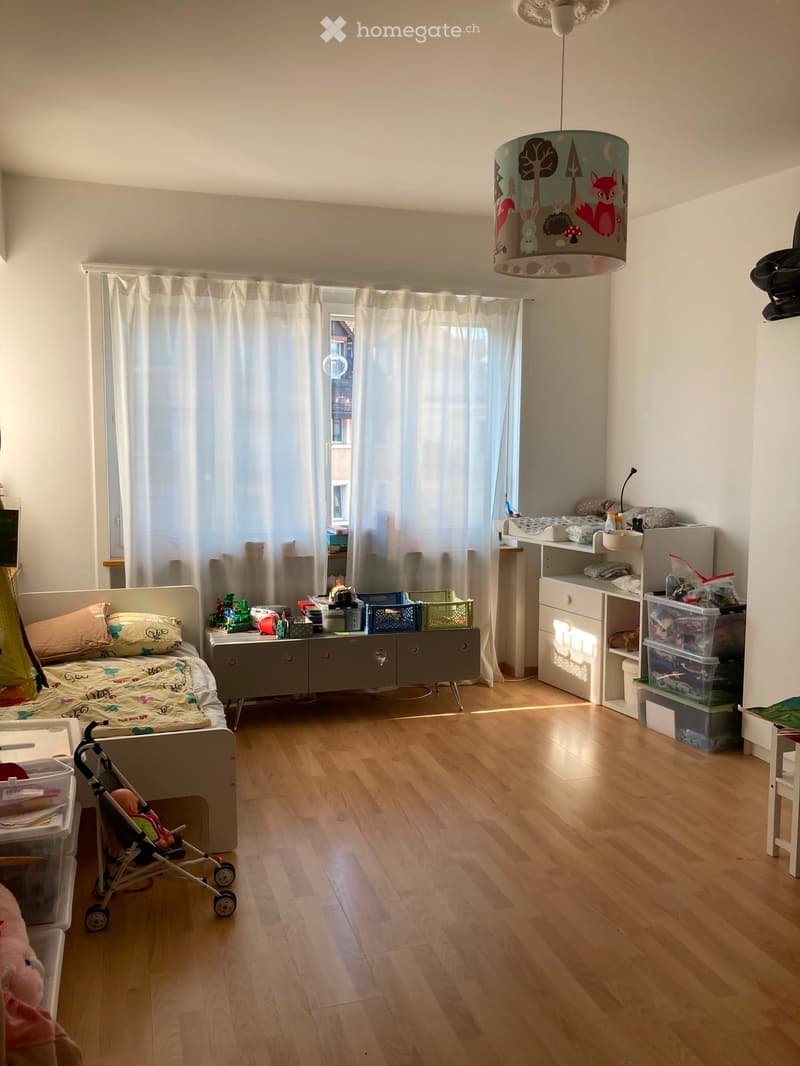 Child's bedroom with a single bed, nightstand, storage unit, and various toys. The room features wooden flooring, large windows with white curtains, and a hanging lamp with a nature-themed shade.