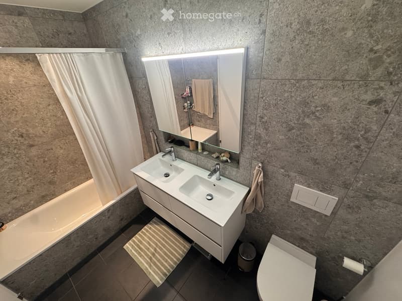 Modern bathroom with a double sink, large mirror, bathtub, and toilet. Features marble walls and a striped bath mat.