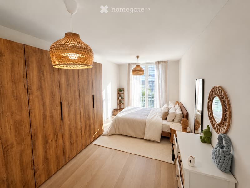 Modern, well-lit bedroom with a large wooden wardrobe, a neatly made bed with pillows and a duvet, a nightstand with a lamp, a round mirror, and decorative items including a woven pendant light and a small plant.