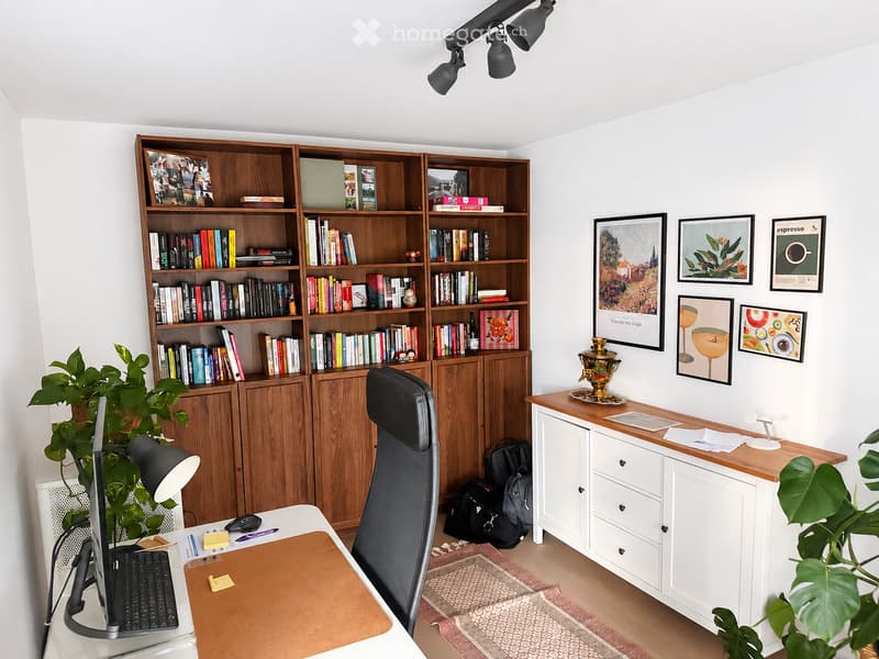 A modern, well-organized home office with a wooden desk, ergonomic chair, bookshelf filled with books, and decorative items.