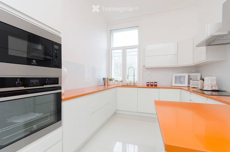 Modern kitchen with white cabinets, orange countertops, stainless steel appliances, and a large window.
