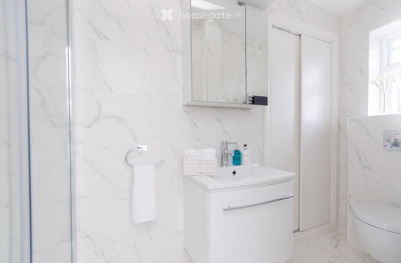 Modern bathroom with white marble walls and floor, a white sink with a cabinet, a mirror above the sink, a towel on a ring, and a toilet in the corner.