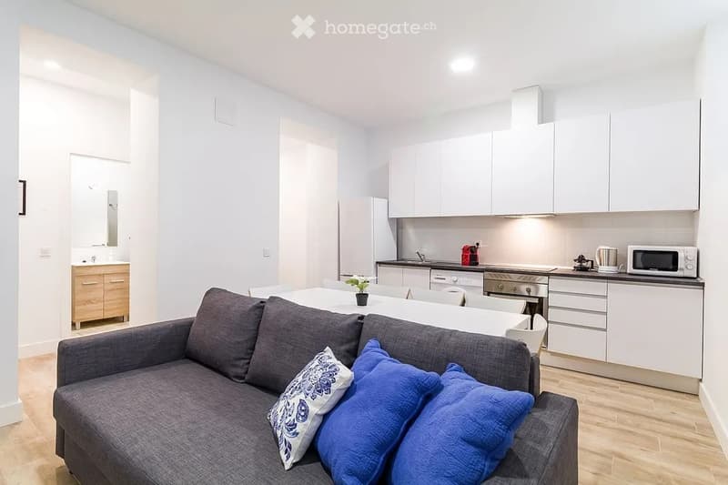 Modern, open-concept living and dining area with a grey sofa, coffee table, and a small dining table. The kitchen features white cabinets, stainless steel appliances, and a microwave on the counter.