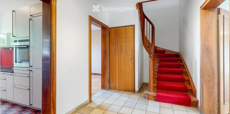 Hallway with a wooden staircase featuring a red carpet, a wooden door, and a view into a modern kitchen with stainless steel appliances.
