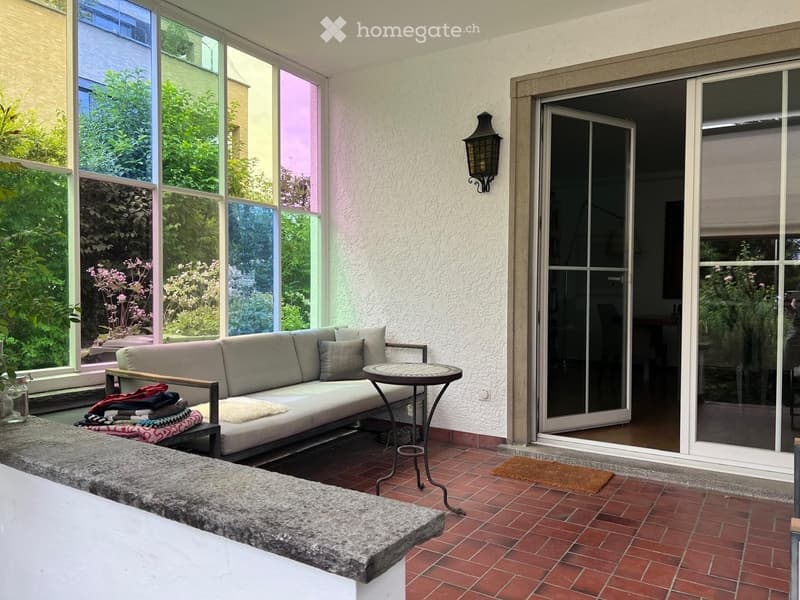 A cozy, sunlit living space with a grey sofa, a small round table, a bench with folded blankets, and a wall-mounted lamp. Large windows offer a view of greenery and a sliding glass door leads to another room.