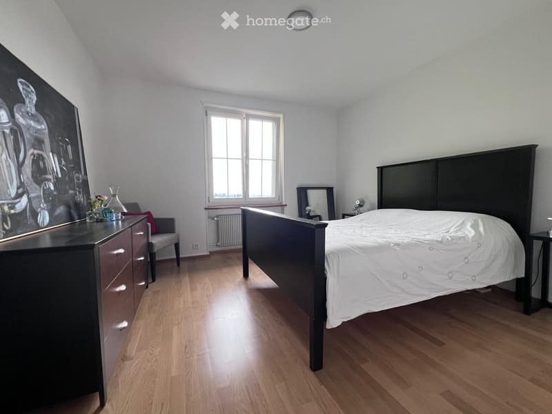 Modern bedroom with wooden flooring, large window, bed with dark headboard, dresser with mirror, armchair, and decorative items on the dresser.