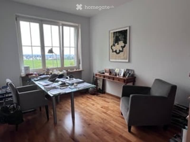 Bright, modern room with large windows, wooden floor, desk with laptop, chair, and books, and a comfortable sofa.