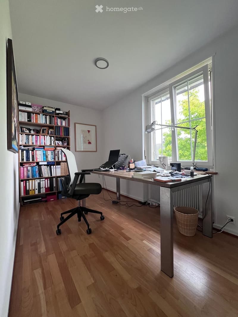 Well-lit office with wooden flooring, large desk, ergonomic chair, bookshelf filled with books, laptop, and a floor lamp near the window.