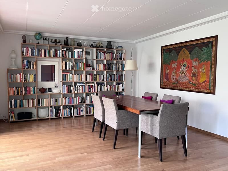 Dining area with a wooden table, six chairs, a large bookshelf filled with books and decorative items, a floor lamp, and a colorful tapestry on the wall.