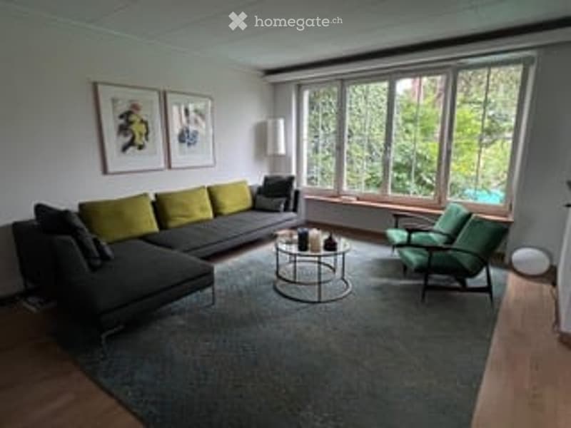 Modern living room with large windows, grey sectional sofa, green armchairs, coffee table, and artwork on the wall.