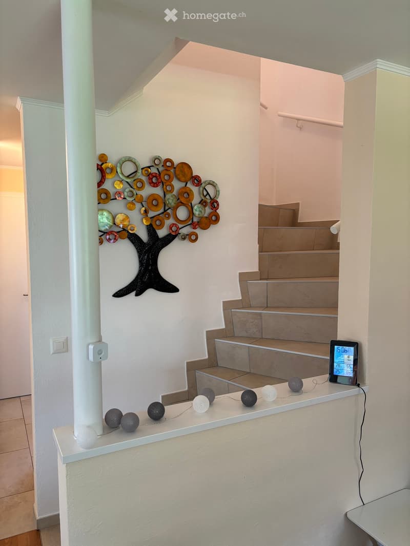 Hallway with a staircase leading upwards, featuring a decorative wall art piece shaped like a tree with colorful circular elements, and a small shelf with spherical decorative items and a digital clock displaying the time and temperature.