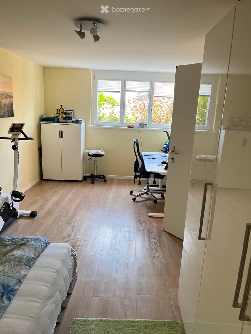 Well-lit room with wooden flooring, a bed with a blue blanket, a desk with a chair, a laptop, a cabinet, and an exercise bike.