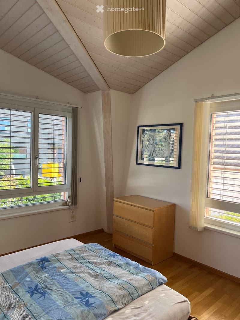 Bright bedroom with wooden floors, large windows with blinds, a bed with blue and white bedding, a wooden nightstand, and a framed picture on the wall.