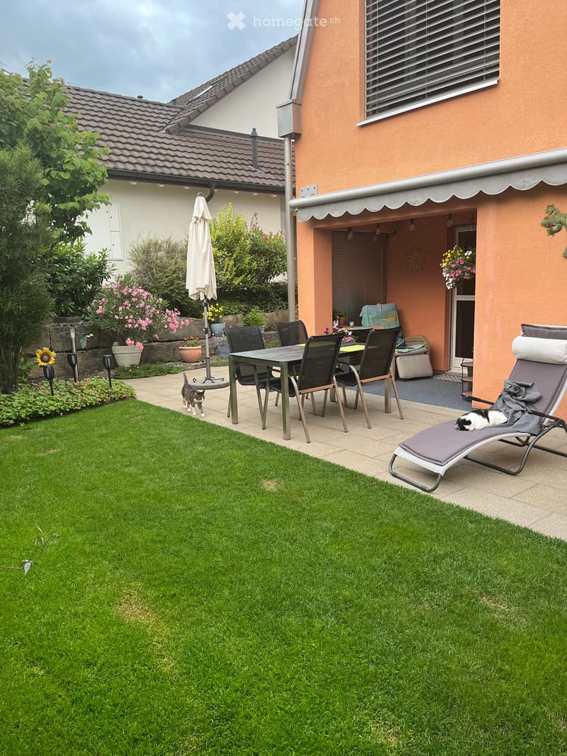 Garden with a well-maintained lawn, patio area with a table, chairs, umbrella, and sun lounger. A cat is seen near the patio.