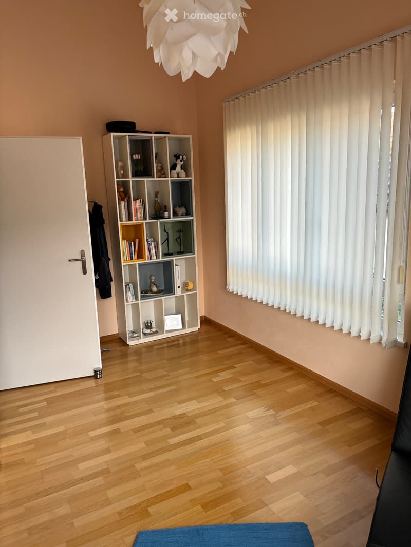 A well-lit room with wooden flooring, a white shelf filled with books and decorative items, a large window with vertical blinds, and a modern white chandelier hanging from the ceiling.