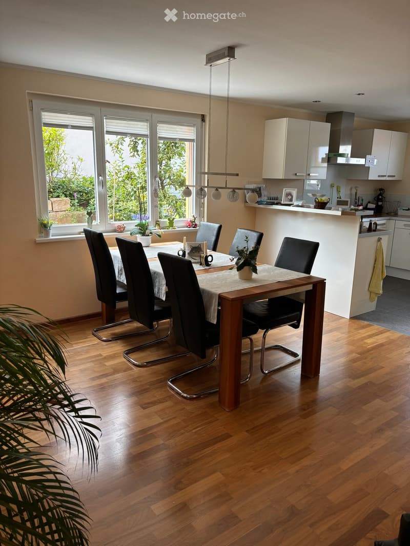 Dining area with a wooden dining table, four black chairs, and a modern kitchen area with white cabinets and a countertop. The room features wooden flooring, large windows with blinds, and a pendant light fixture.