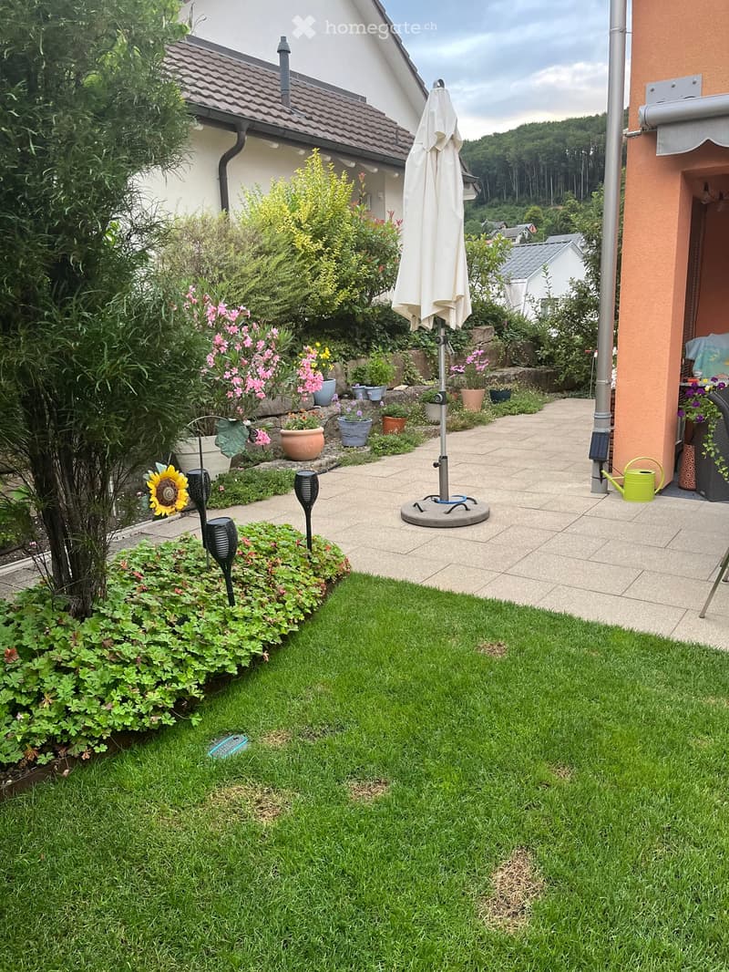 Well-maintained garden with a lawn, flower beds, potted plants, and a patio area with an umbrella and outdoor lighting.