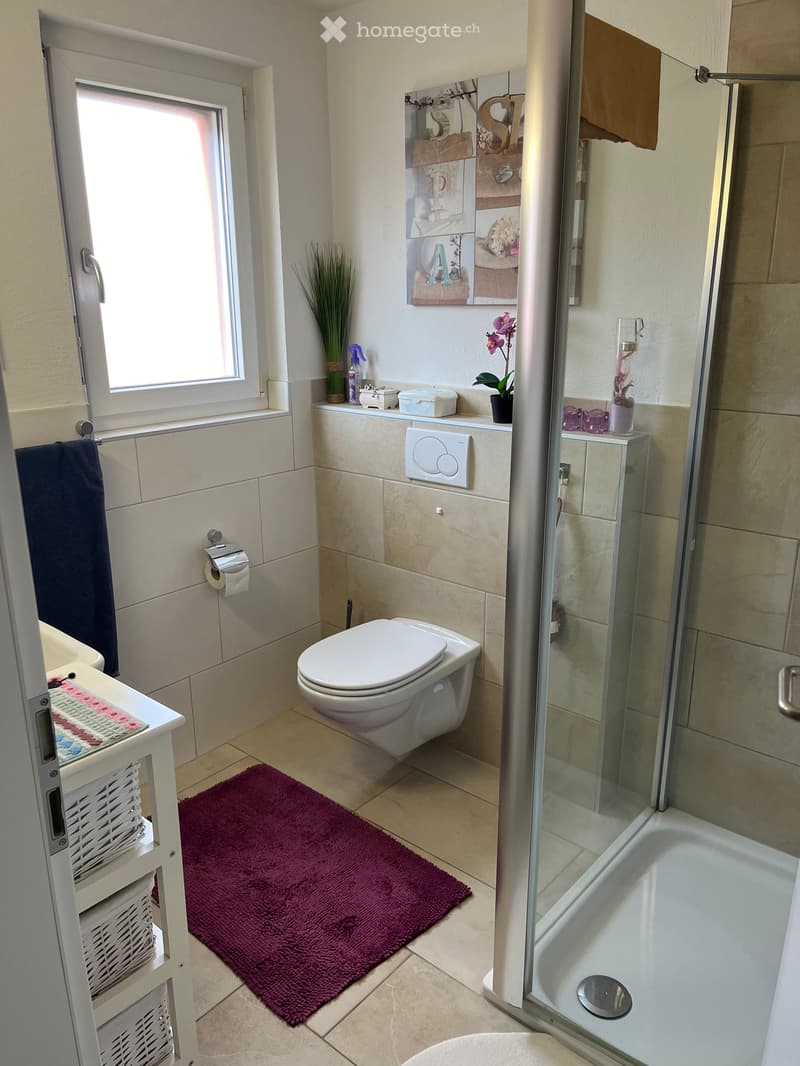 Modern bathroom with a toilet, bathtub, glass shower enclosure, and a small shelf with decorative items. Features a purple bath mat, a white basket, and a window with a view.