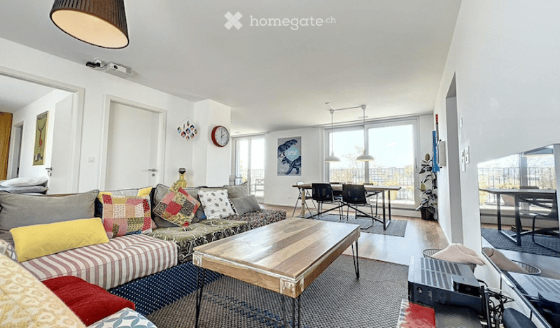 Bright, open-plan living and dining area with large windows and balcony access. Features a patterned sofa with colorful cushions, a wooden coffee table, dining chairs and table, and modern lighting.