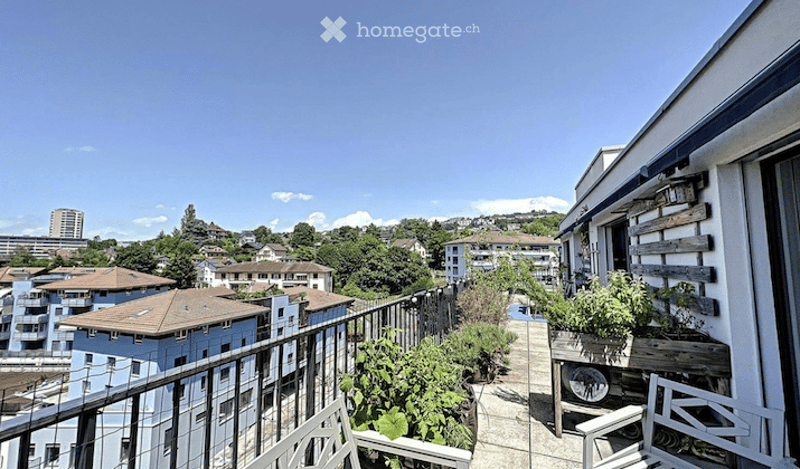Balcony with metal railing, wooden decking, potted plants, and a view of the city and surrounding buildings.