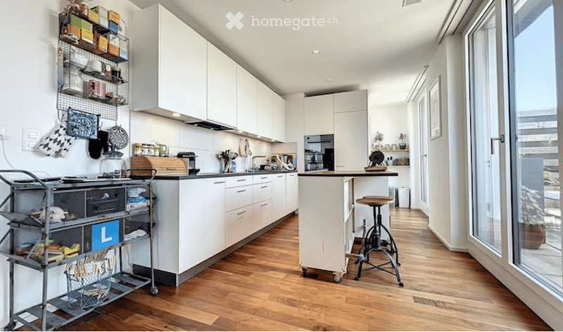 Modern kitchen with white cabinets, wooden flooring, and various appliances including a sink, stove, and microwave. A small island with a stool is present, along with a cart holding various items.