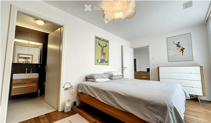 Modern bedroom with a wooden bed, nightstand, and a small adjacent bathroom with a sink and mirror. The room features a framed picture and a poster on the wall, along with a hanging lamp.