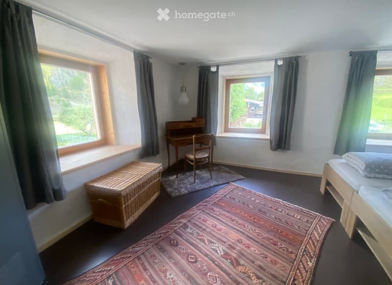 Cozy bedroom with a bed, desk, chair, wicker chest, and patterned rug. Large windows with curtains provide natural light.