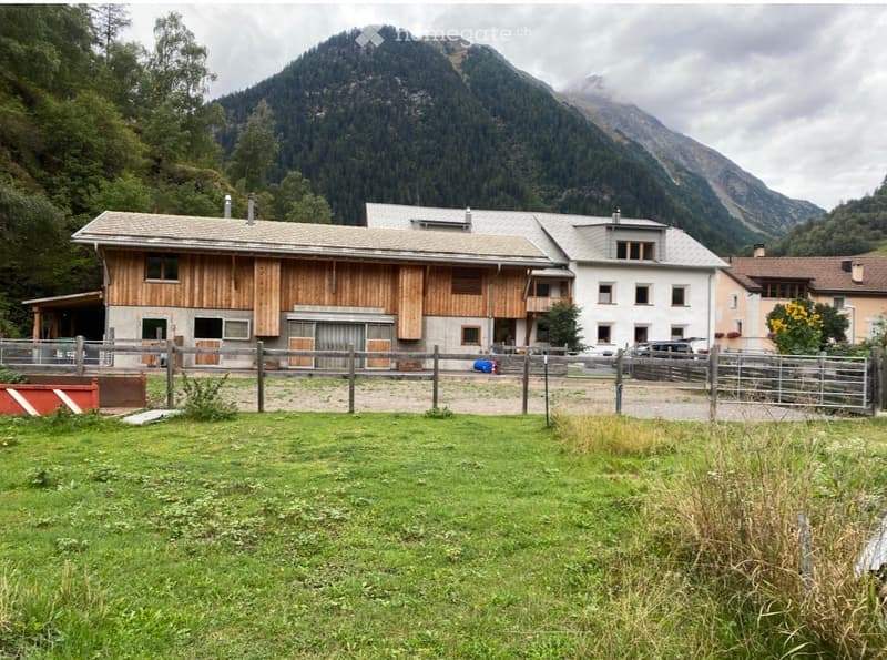 A multi-building complex with a wooden stable, a white house, and a fenced yard, set against a mountainous backdrop.