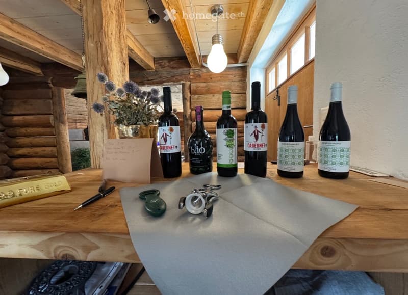 A wooden table with six wine bottles, a pen, a bottle opener, and a decorative flower vase. The background features exposed wooden beams and a hanging light fixture.