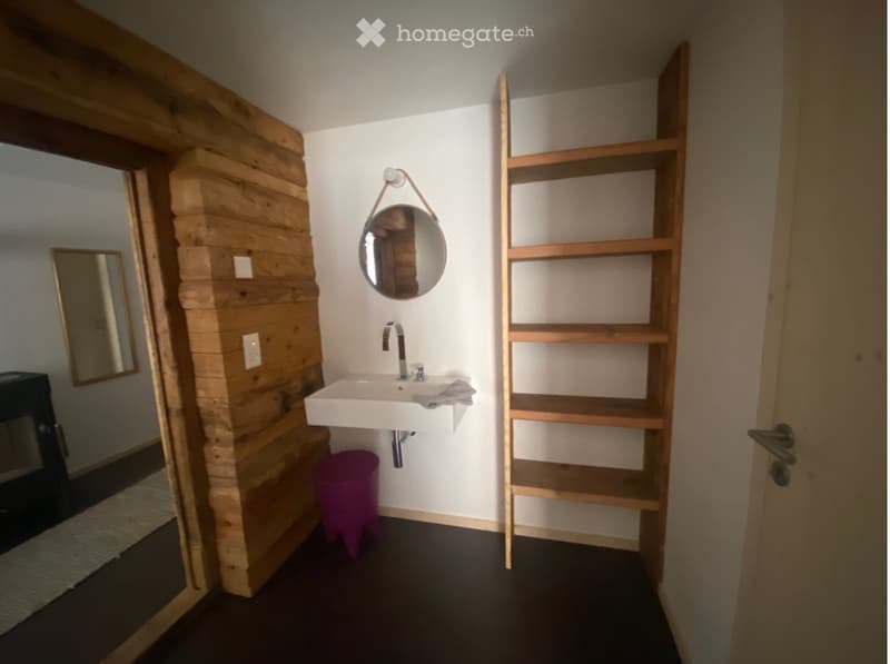 Modern bathroom with white sink, round mirror, wooden shelf, and purple stool. Wooden wall paneling and dark flooring.