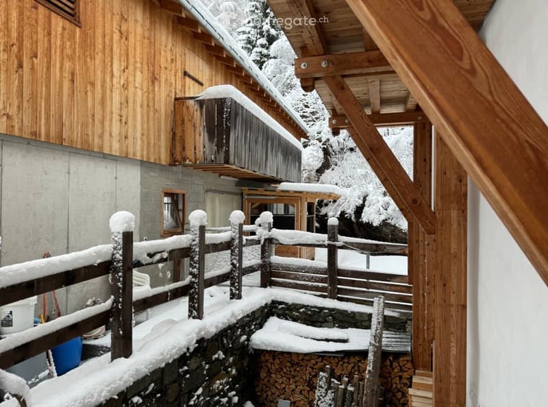Snow-covered wooden deck and stone steps leading to a wooden fence, with a stack of firewood and a small shed in the background.