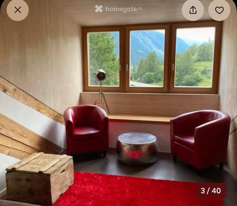 A cozy, modern living space with red chairs, a small round table, a floor lamp, and a wooden chest, overlooking a scenic mountain view.