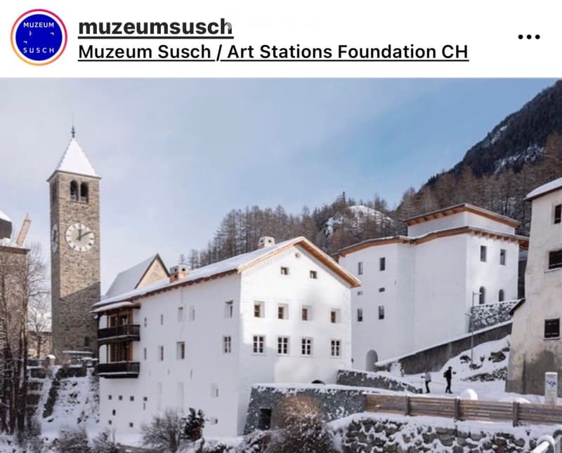 A historic white building complex with a tall clock tower, surrounded by snow-covered ground and trees, with a mountainous backdrop.