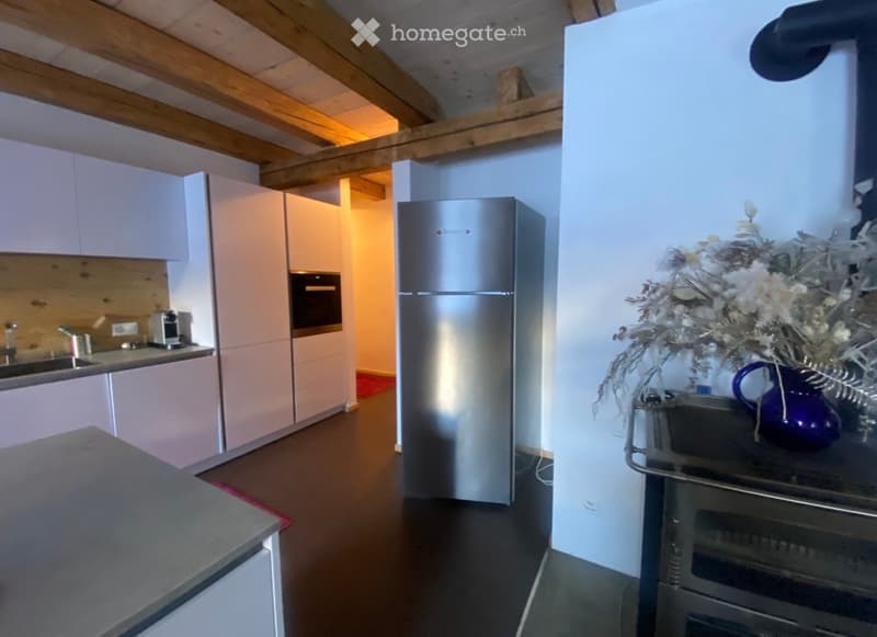 Modern kitchen with light-colored cabinets, stainless steel appliances, and a wooden ceiling beam. Features include a refrigerator, oven, sink, coffee maker, and a decorative vase with flowers on the counter.