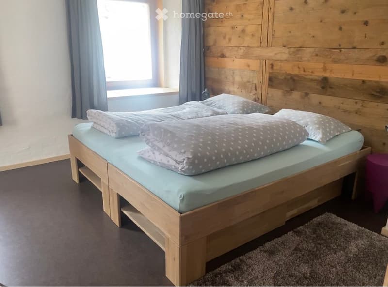 Cozy bedroom with a wooden bed, light blue bedding, and a grey rug on the floor. The room features wooden panel walls and a window with blue curtains.