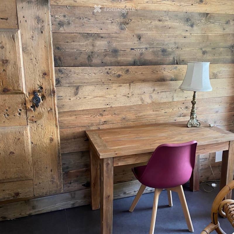 A small, furnished room with a wooden desk, a pink chair, and a table lamp on the desk. The walls are made of reclaimed wood planks, and the floor is dark blue.