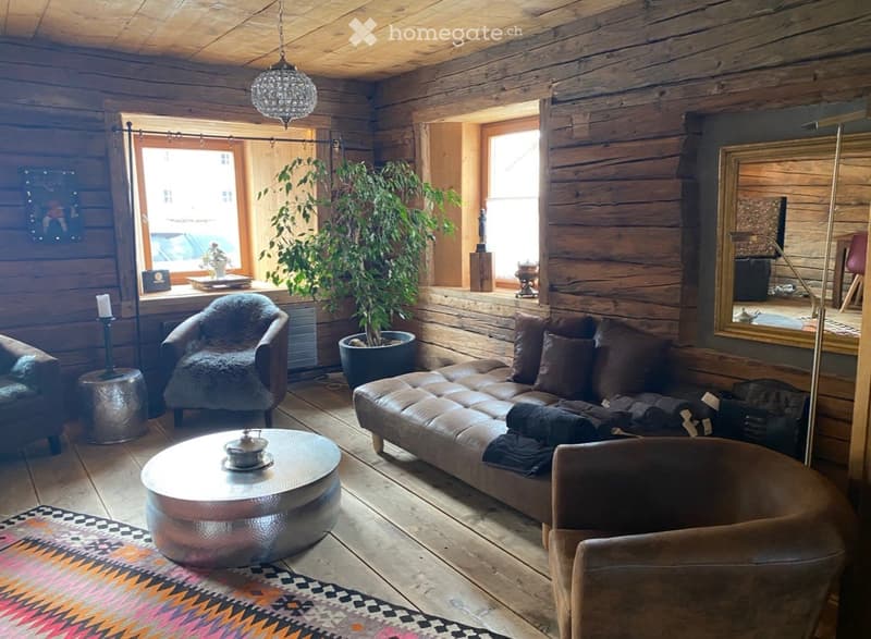 Cozy living room with wooden walls and floors, a large sofa, coffee table, armchair, floor lamp, and a potted plant near the window.