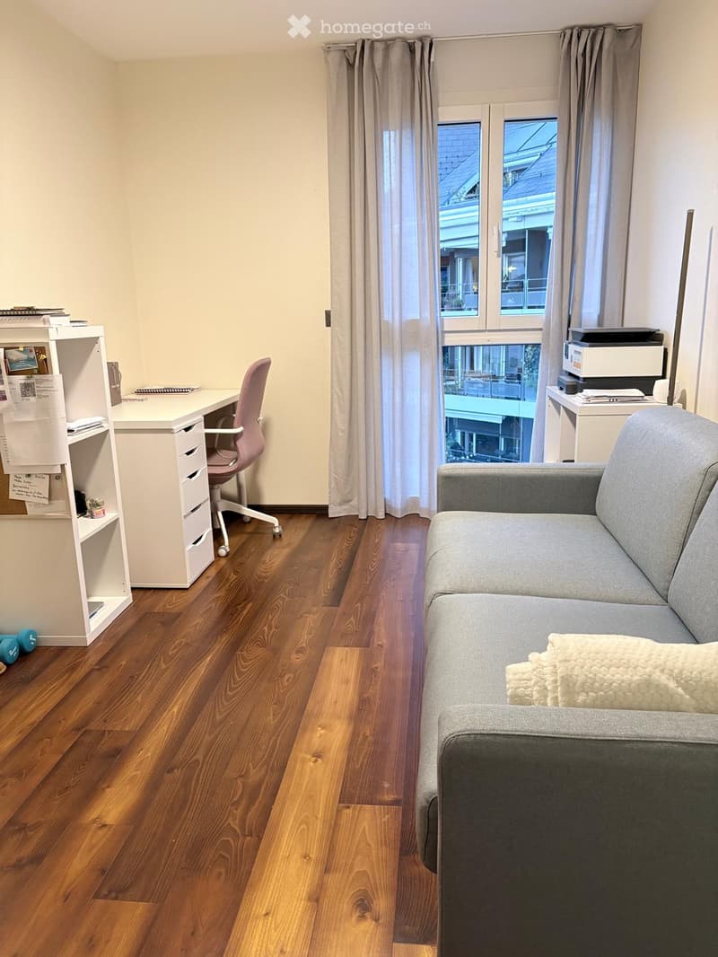 A modern living room with a grey sofa, a pink chair, a white desk, and a shelf with books and papers. The room features wooden flooring and large windows with curtains.