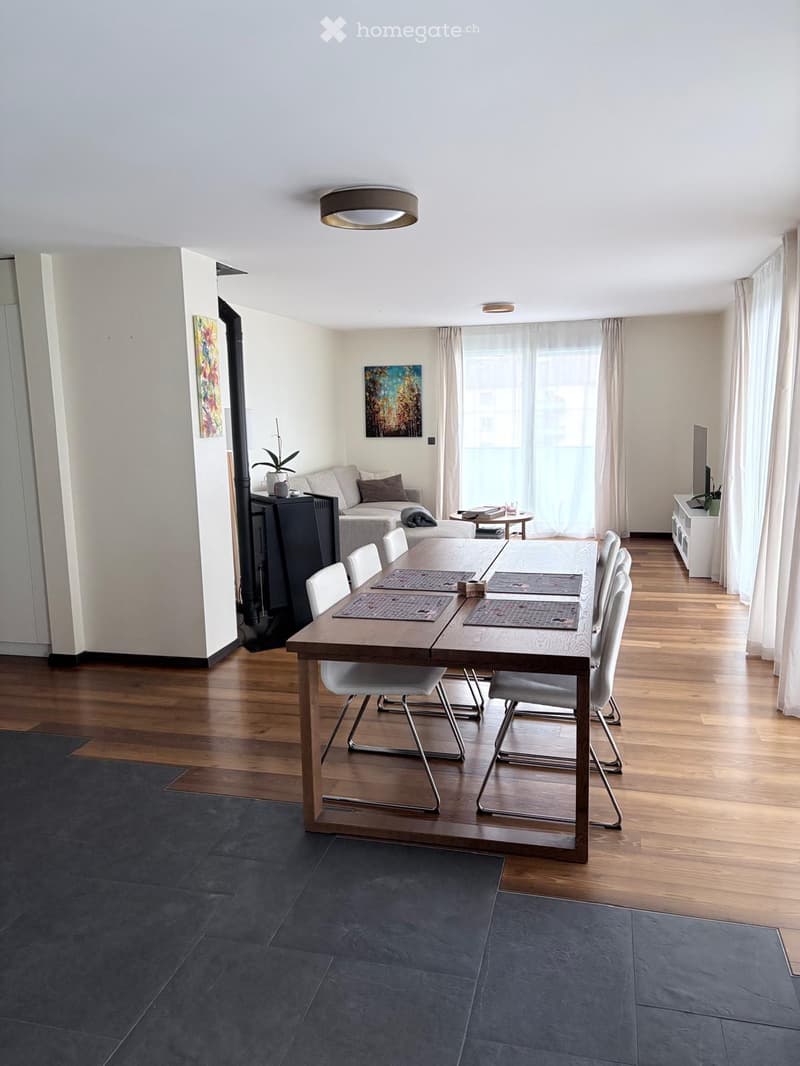 Modern open-plan living and dining area with wooden flooring, a large dining table with six chairs, a sofa, a TV, and artwork on the walls.