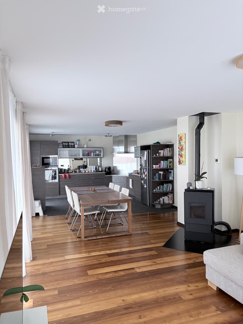 Open-plan living and dining area with wooden flooring, a dining table with chairs, a fireplace, a bookshelf, and modern kitchen appliances including a refrigerator, oven, and microwave.