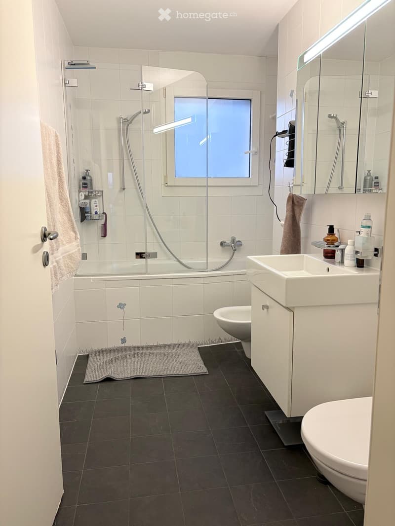 Modern bathroom with white tiles, bathtub, sink, toilet, mirror, and shower area. Features a window, towel, and various bathroom accessories.