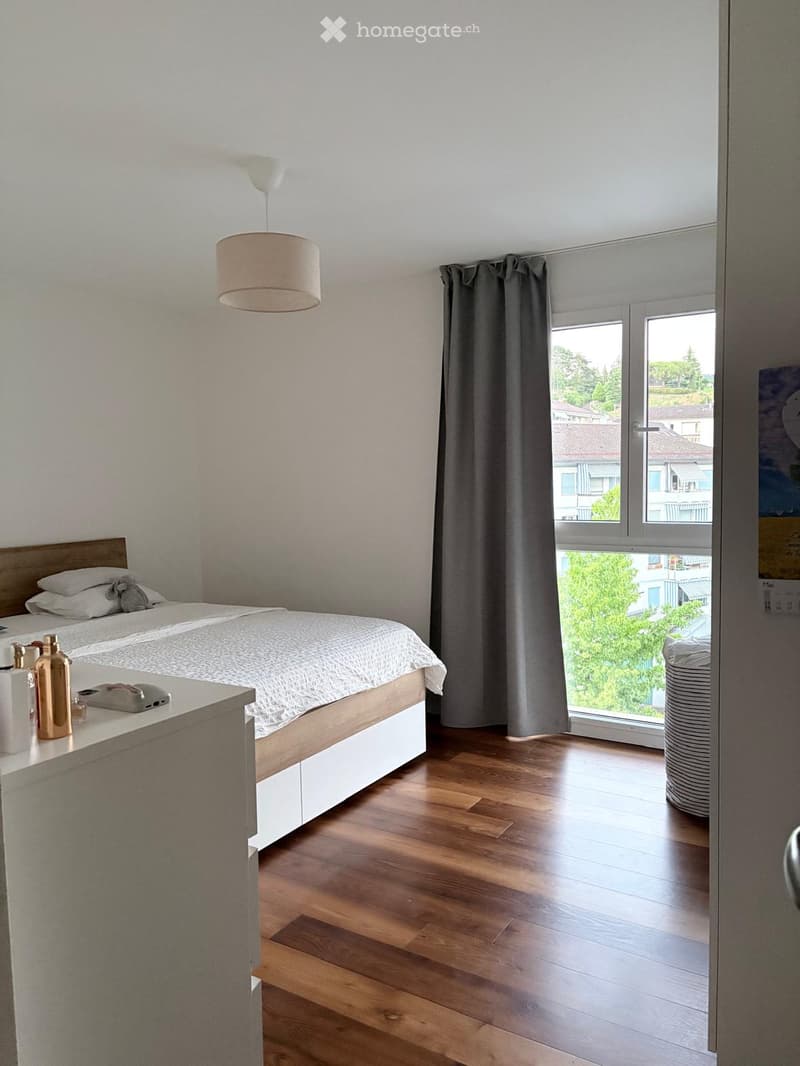 Modern bedroom with a wooden bed, nightstand, lamp, and large window with curtains. The room features wooden flooring and a view of greenery outside.