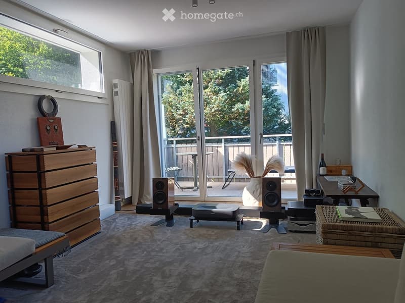 Modern living room with large windows and sliding glass doors leading to a balcony. Features a grey sofa, wooden cabinet, speakers, and decorative items.