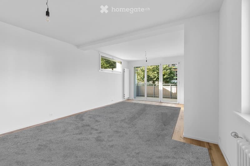 Empty room with white walls, wooden flooring, grey carpet, large window, and sliding glass doors leading to a balcony.