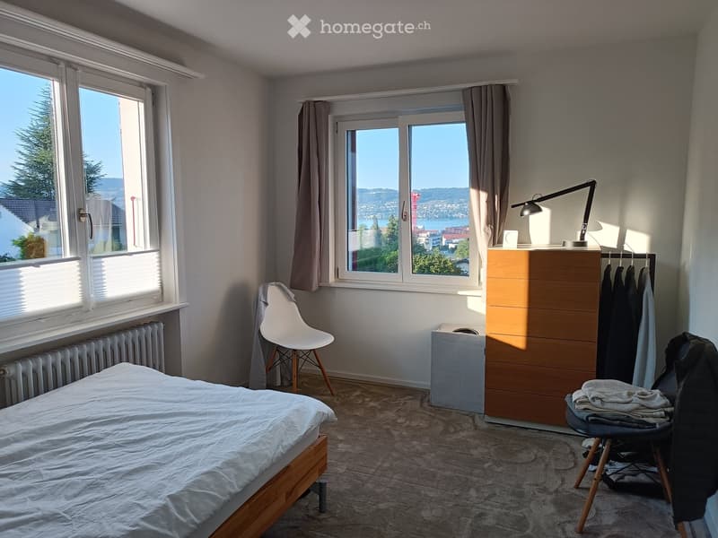 Bright bedroom with large windows offering a scenic view, featuring a bed, a white chair, a wooden wardrobe, a desk lamp, and a radiator.