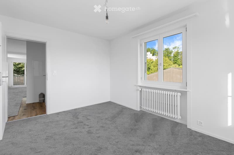 Empty room with white walls, grey carpet, large windows, radiator, and a door leading to another area.