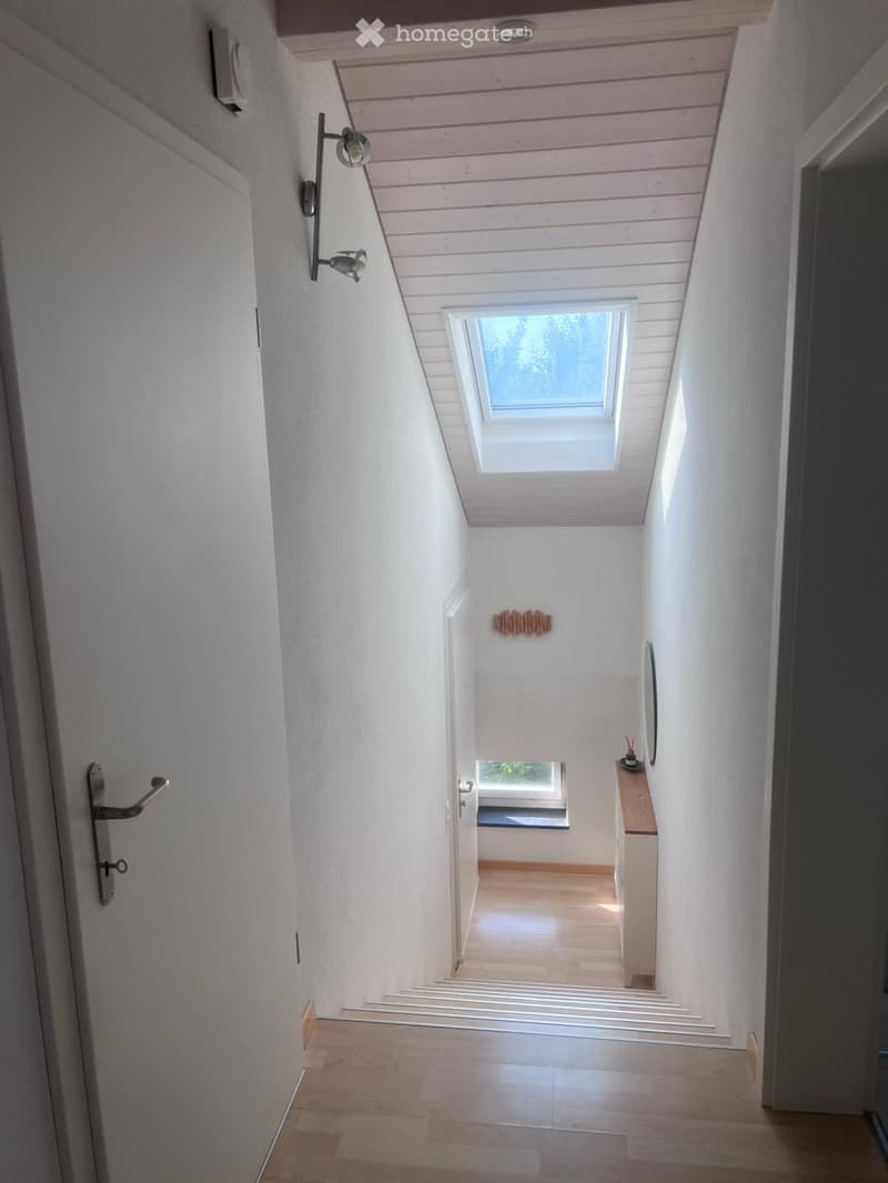 Modern hallway with wooden ceiling, white walls, and a skylight window at the top. Features a small wooden shelf with decorative items and a small table with a lamp.