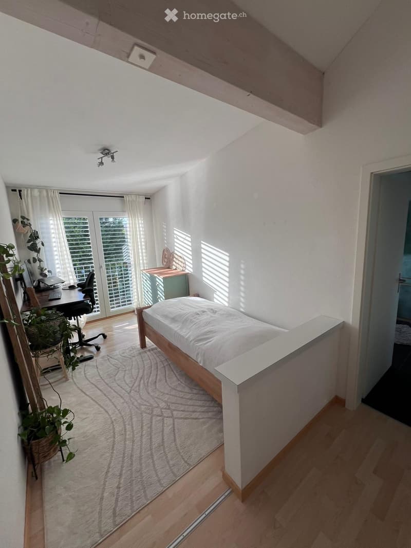 Bright bedroom with a wooden bed, desk, chair, laptop, and a plant near the window. The room features white walls, wooden flooring, and large windows with blinds.